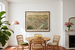 A large framed vintage map print of a city hangs on a white wall in a styled dining room with a round wooden table, four wicker chairs, a pink floor lamp, and a fireplace with flowers.