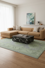 A modern living room scene featuring a tan sectional sofa, a black marble coffee table, and a large sage green patterned area rug on a light wood floor. A white wall in the background has a shelf with decorative vases and a framed abstract print.