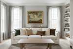 A styled living room featuring a white sofa with beige and brown pillows, a wooden coffee table, gray curtains, and a framed landscape painting on the wall.