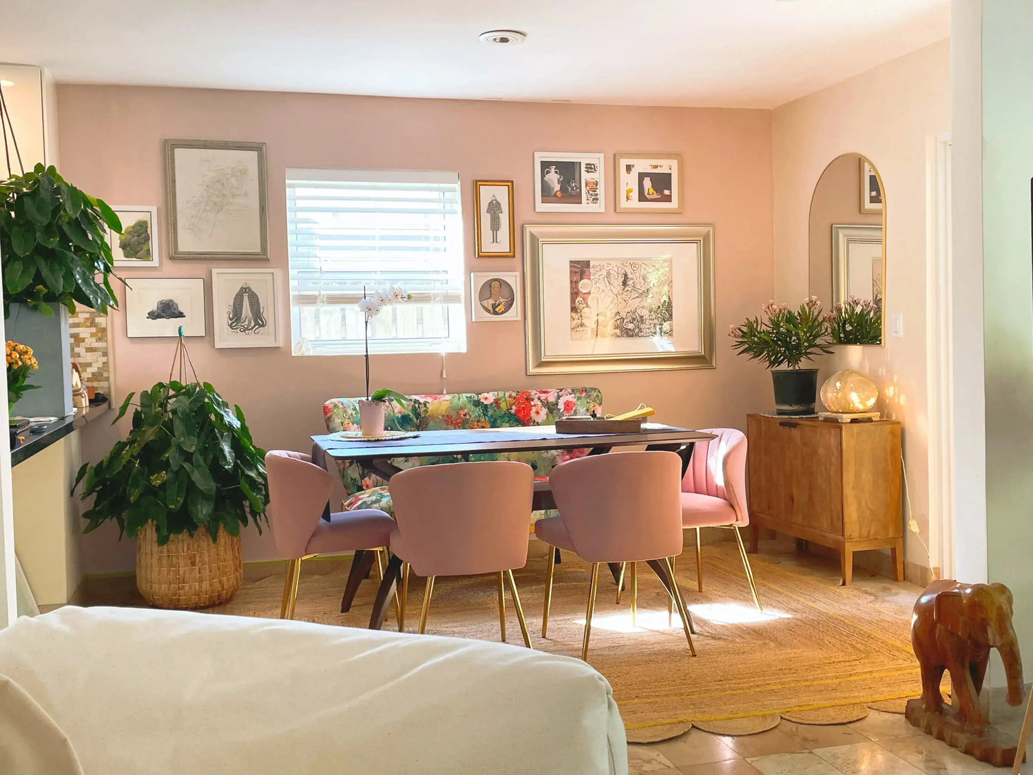A cozy room featuring a Handmade Natural Color Scalloped Border Jute Rug, perfect as an area rug in an office or bedroom. The space is decorated with vibrant pink chairs and a floral centerpiece, complemented by a gallery wall and lush greenery, creating a warm and inviting atmosphere.