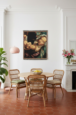A framed classical painting hangs on a white wall above a round wooden dining table and four woven chairs in a bright, modern dining room with terracotta hexagonal tile flooring.