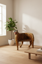 A tiger-shaped decorative sculpture stands on a light wood floor in a bright room with cream walls, a potted plant, and a low wooden table.