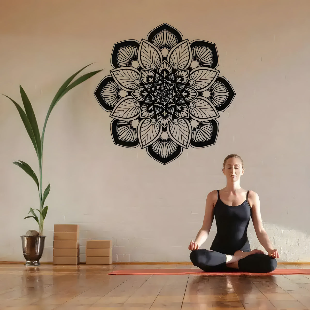 Black mandala flower metal wall art on beige brick wall above a meditating woman and potted plant