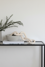 A modern beige chunky chain-link sculpture draped over a matching beige ceramic bowl, styled on a glass-topped console table with a potted plant in the background.