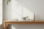 A minimalist interior scene with a light wood console table against a white wall, styled with stacked books, a white bowl, a sculptural cream chain, and a tall cream vase. Sunlight casts soft shadows from a hanging plant onto the wall.
