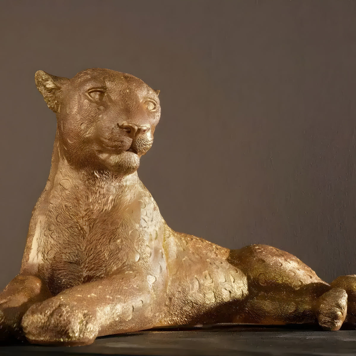 Gold reclining leopard sculpture, head raised and front paws crossed, on dark backdrop