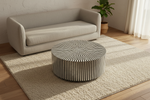 A round coffee table with a black and white striped bone-slat pattern sits on a beige area rug in a modern living room setting next to a light beige sofa.