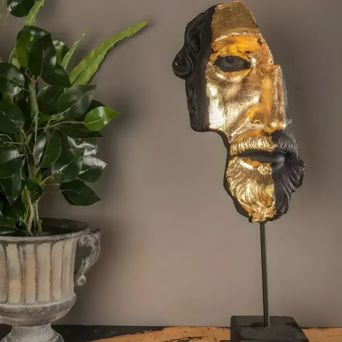 Half gold-gilded, half-black bearded Zeus face sculpture on a metal stand beside a potted plant