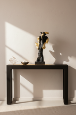 A black console table against a warm beige wall, styled with a dark and gold abstract sculpture, a small bowl, and a dried branch under dramatic diagonal sunlight casting shadows.