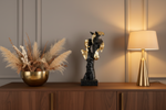 A decorative black horse sculpture with metallic gold accents sits on a wooden console table in a warmly lit room, flanked by a gold bowl with dried grasses on the left and a lit brass table lamp on the right.