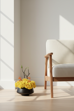 A black ceramic planter filled with bright yellow flowers and small orange blossoms, placed on a light wood floor next to a modern armchair in a sunlit room.