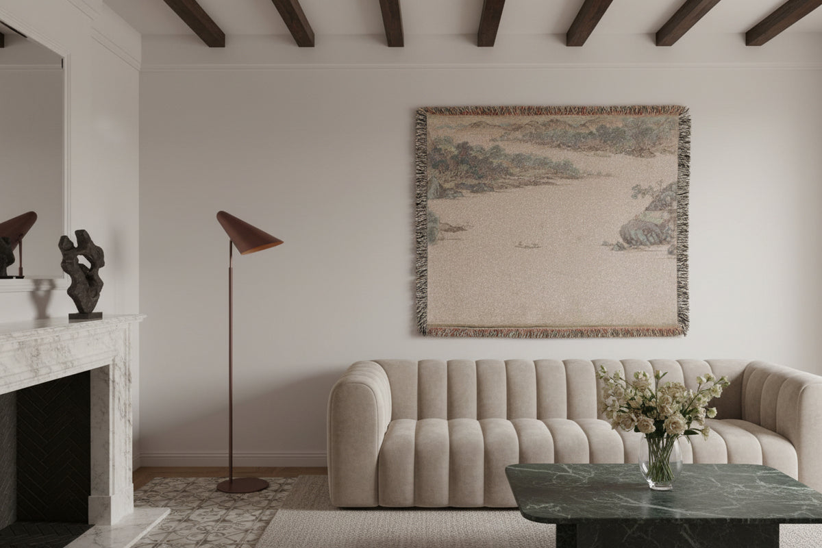 A rectangular woven throw blanket with fringed edges, featuring a muted landscape print in beige, gray, and brown tones depicting trees, rocks, and water.