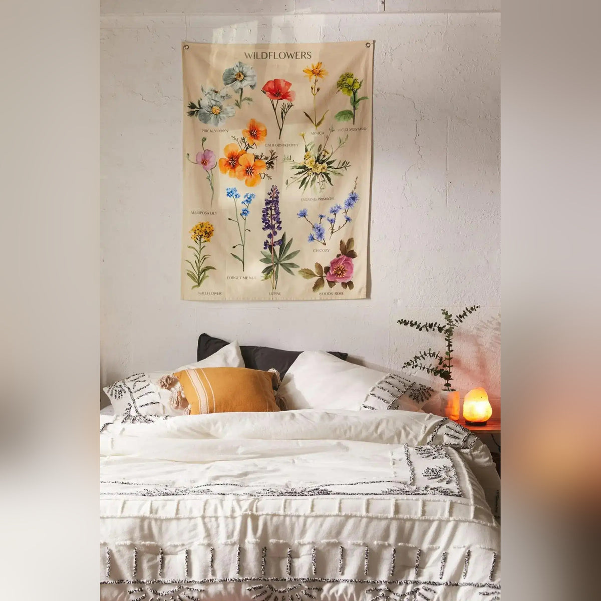Botanical wildflower tapestry on white wall above styled bed — cream fabric with colorful labeled flower illustrations