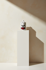 A small white cylindrical container with a lid, featuring a stylized face with black eyes and bright red lips, displayed on a white rectangular pedestal against a beige wall with dramatic shadows.