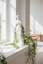 A white classical-style female statue stands on a sunlit windowsill, surrounded by trailing green ivy in a bright, modern interior with white walls and soft natural light.