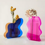 Blue and pink wavy-edged art deco acrylic vases holding dried flowers, translucent curved layered panels on white surface