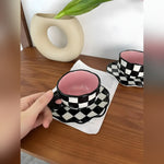 Two checkerboard ceramic mugs with pink interior and wavy saucers on wood table