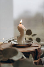 Female torso wax candle lit and glowing on a silver dish atop a wood slice, with soft bokeh eucalyptus leaves in foreground