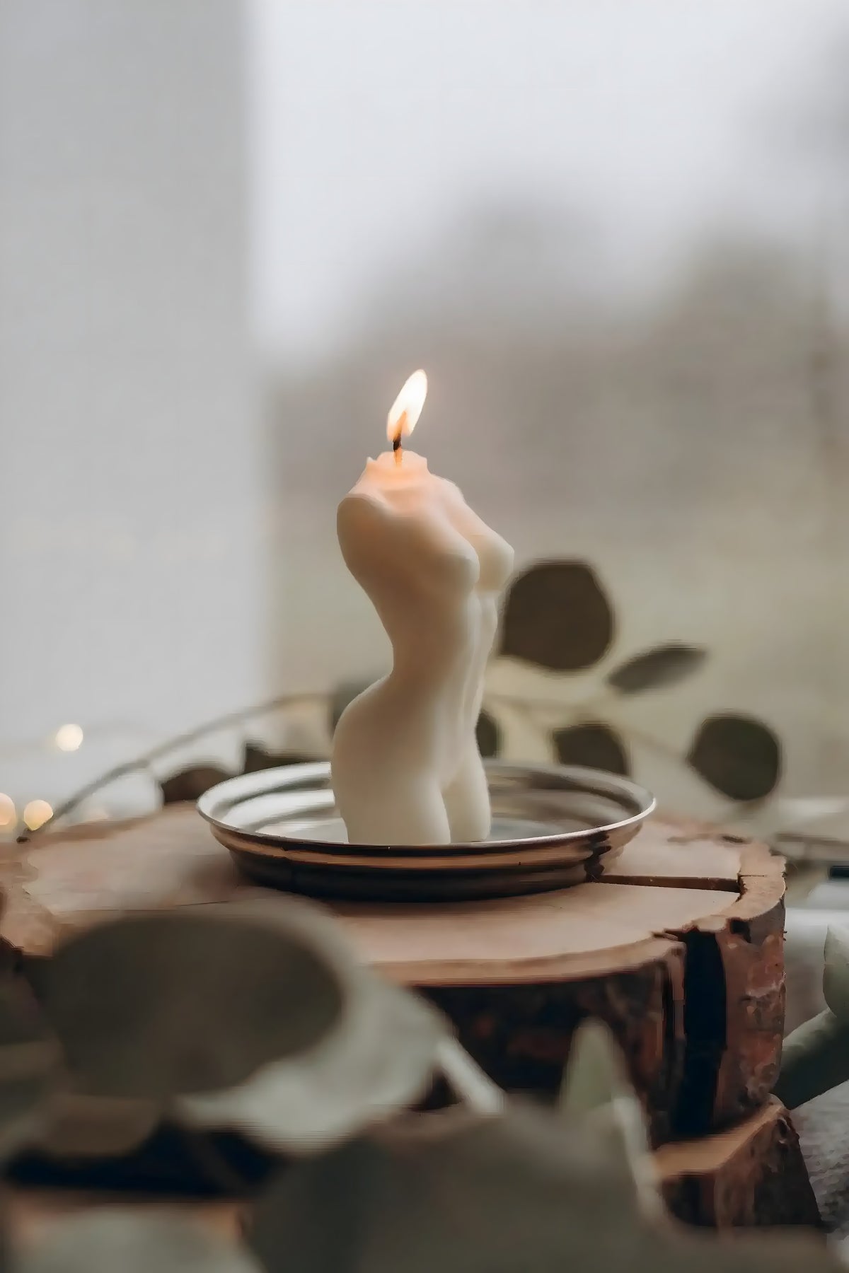 Female torso wax candle lit and glowing on a silver dish atop a wood slice, with soft bokeh eucalyptus leaves in foreground