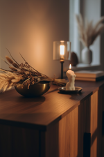 A warm, cozy interior scene showing a wooden sideboard with a brass bowl, dried pampas grass, a lit candle holder, and a white candle, illuminated by soft ambient light.