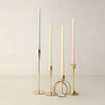 Four slim taper candles in light blue, mint green, cream, and mauve in brass holders on neutral background