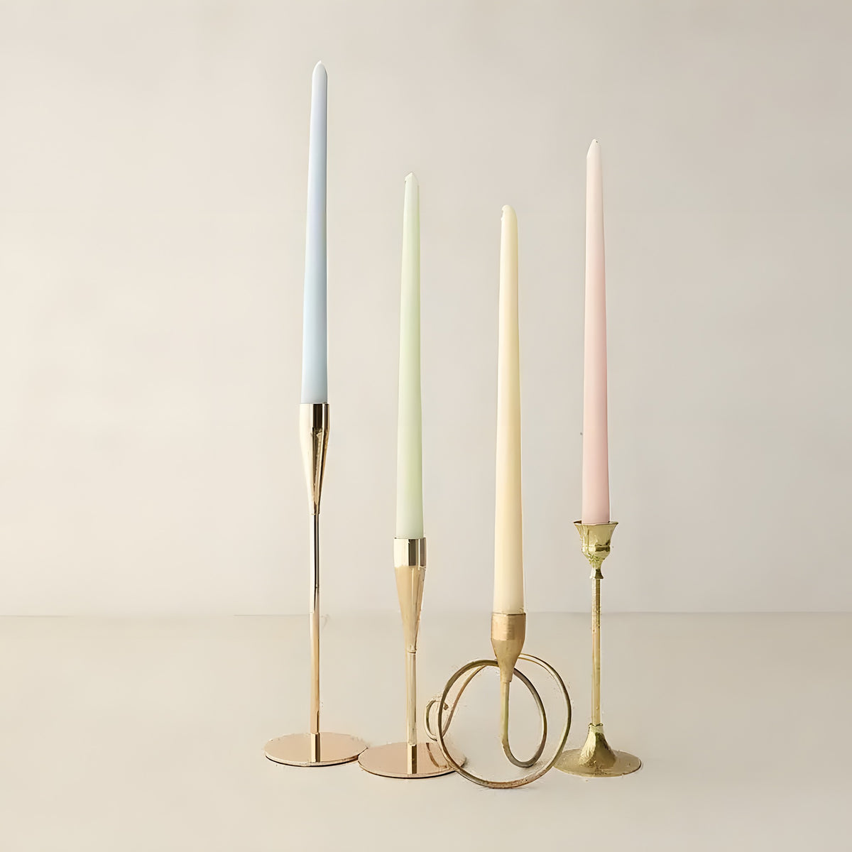 Four slim taper candles in light blue, mint green, cream, and mauve in brass holders on neutral background