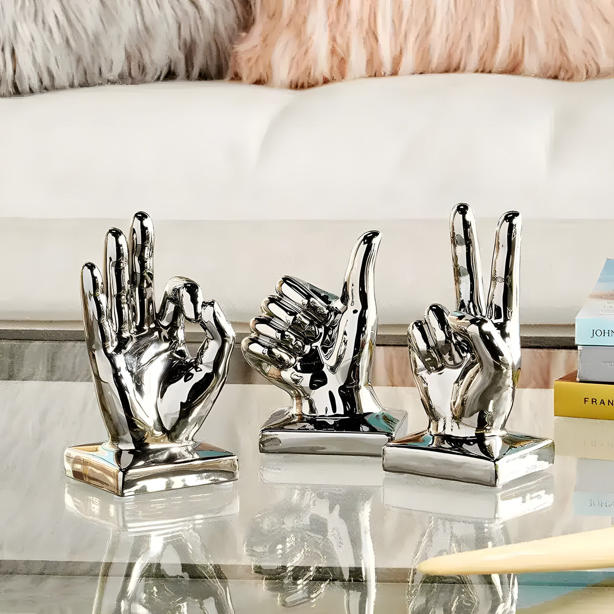 Three polished silver hand sculptures in OK, rock, and peace gestures on glass table before white sofa