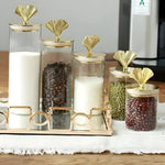 Multiple glass storage jars of varying heights with gold ginkgo-leaf knob lids, filled with pantry goods on a mirrored tray