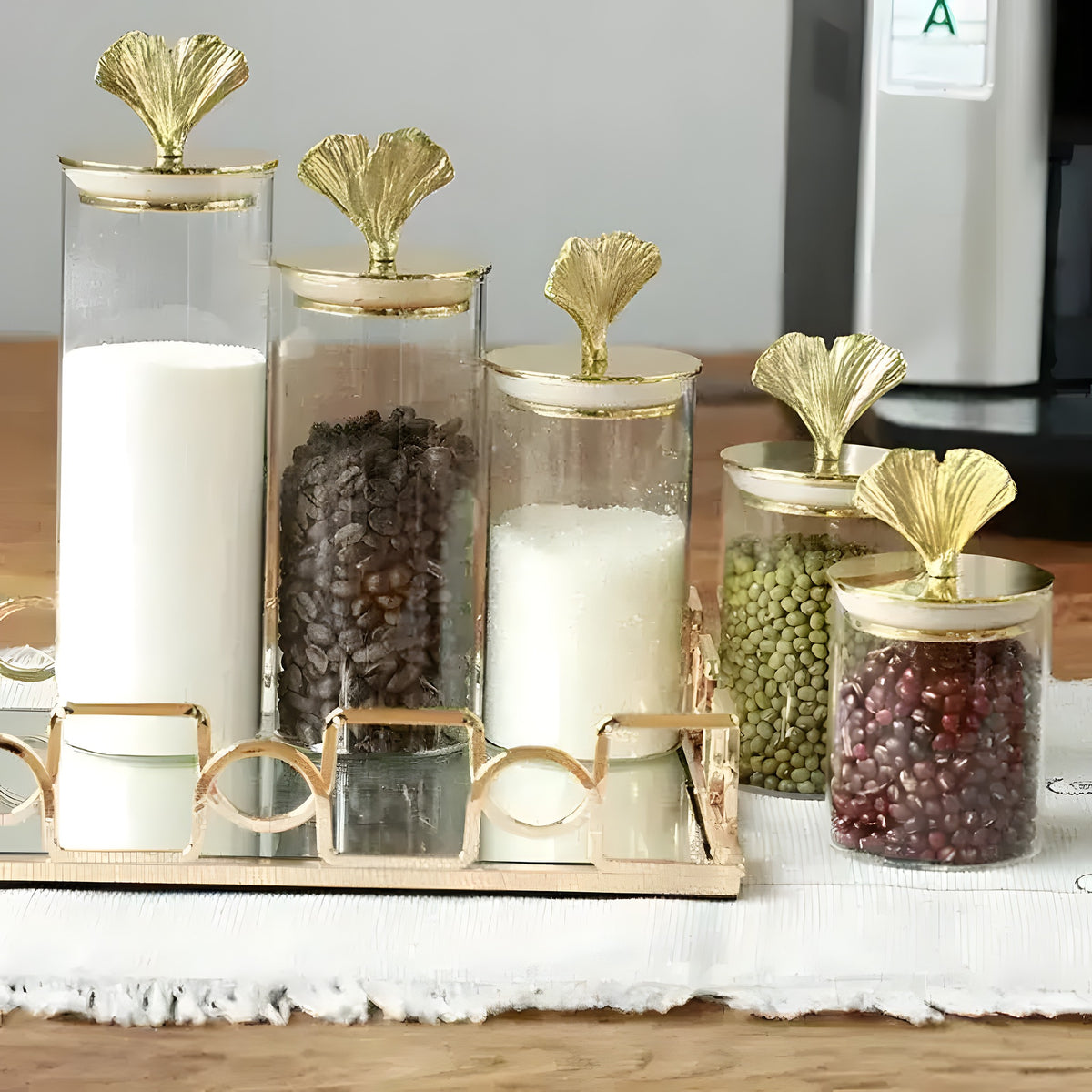 Multiple glass storage jars of varying heights with gold ginkgo-leaf knob lids, filled with pantry goods on a mirrored tray