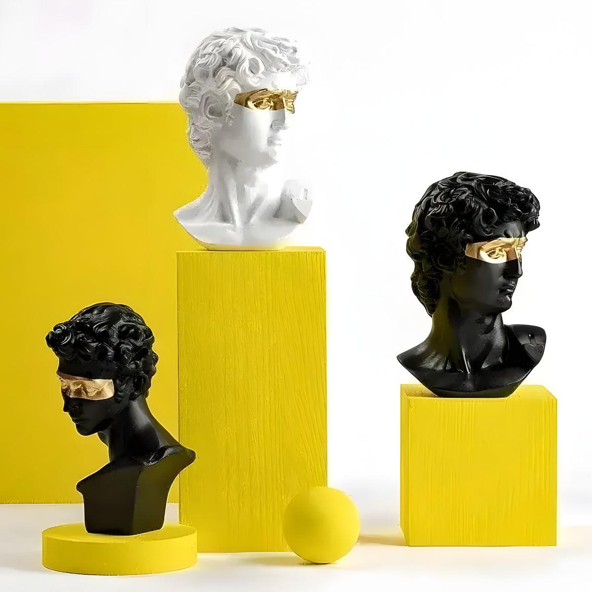 Three David head bust statues with gold eye masks on yellow geometric stands, two black and one white