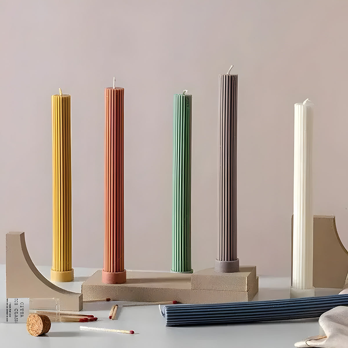 Five tall ribbed column soy candles in yellow, terracotta, green, taupe, and white on concrete geometric stands
