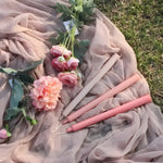 Taupe and dusty pink taper candles laid on draped sheer fabric with pink roses on grass