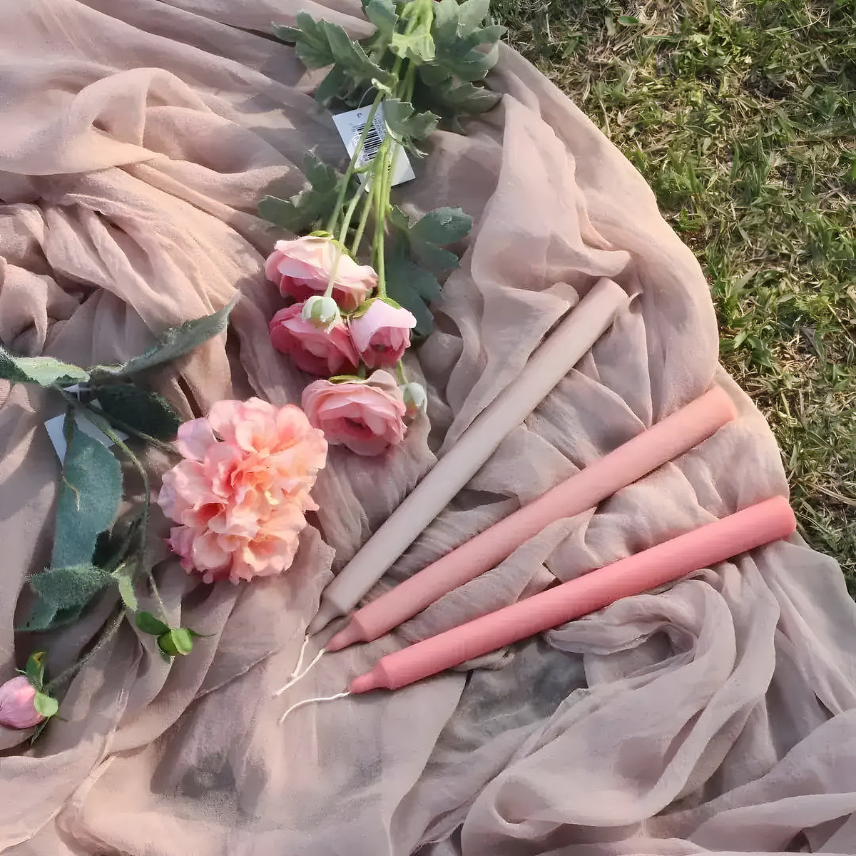 Taupe and dusty pink taper candles laid on draped sheer fabric with pink roses on grass