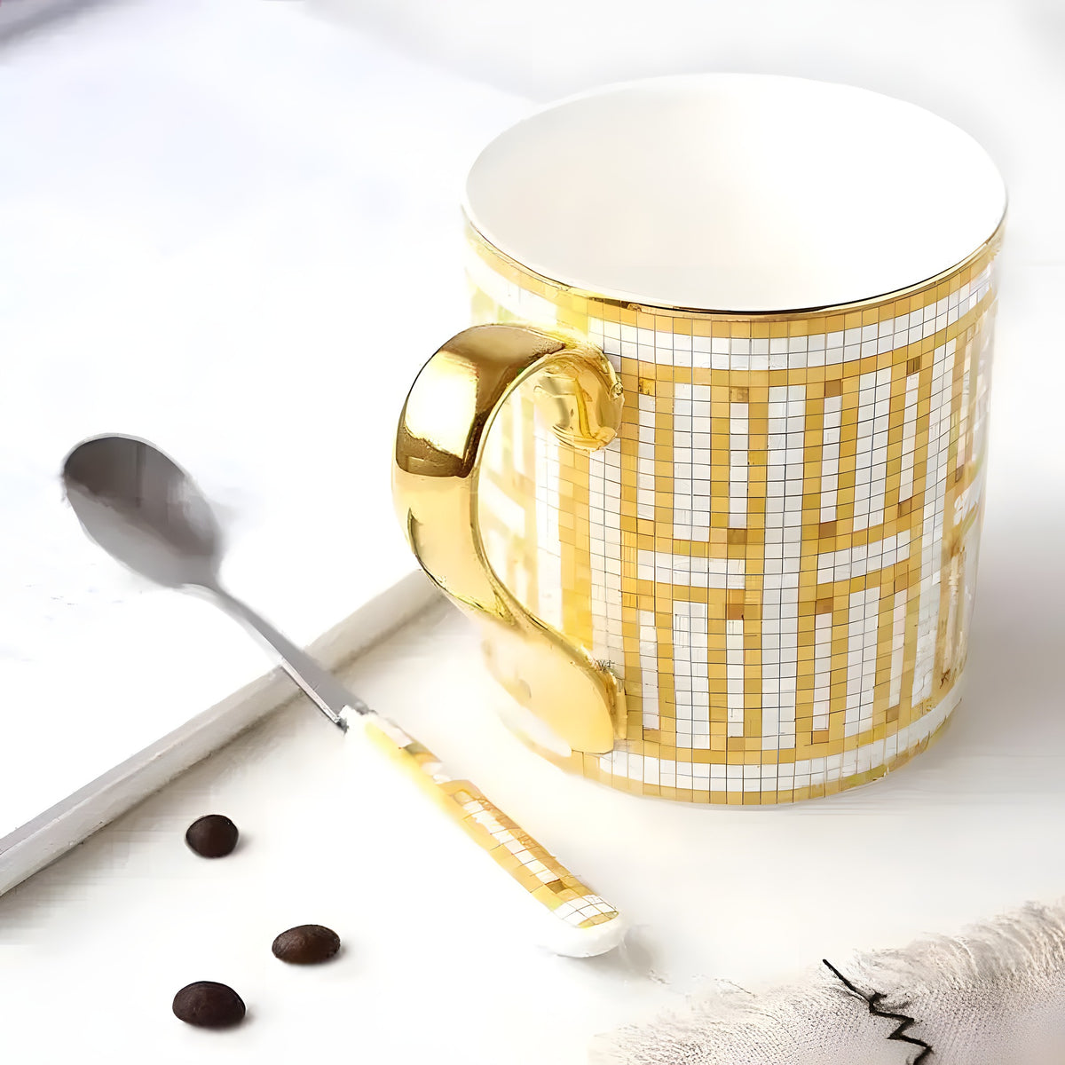 Golden mosaic porcelain coffee mug with gold handle and matching spoon beside a steel spoon and coffee beans on white