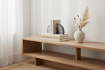 A light natural wood TV stand or console shelf styled with a stack of books, blue agate bookends, and a ceramic vase with dried pampas grass against a white wall and sheer curtain.