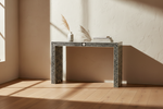 A console table with a black and white chevron bone-inlay pattern, styled with a small vase and books on a light wood floor against a textured beige wall with warm sunlight shadows.