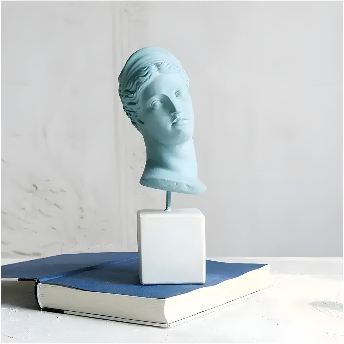 Blue-teal Greek goddess of hunting bust sculpture on white cube pedestal atop a book, textured white wall backdrop