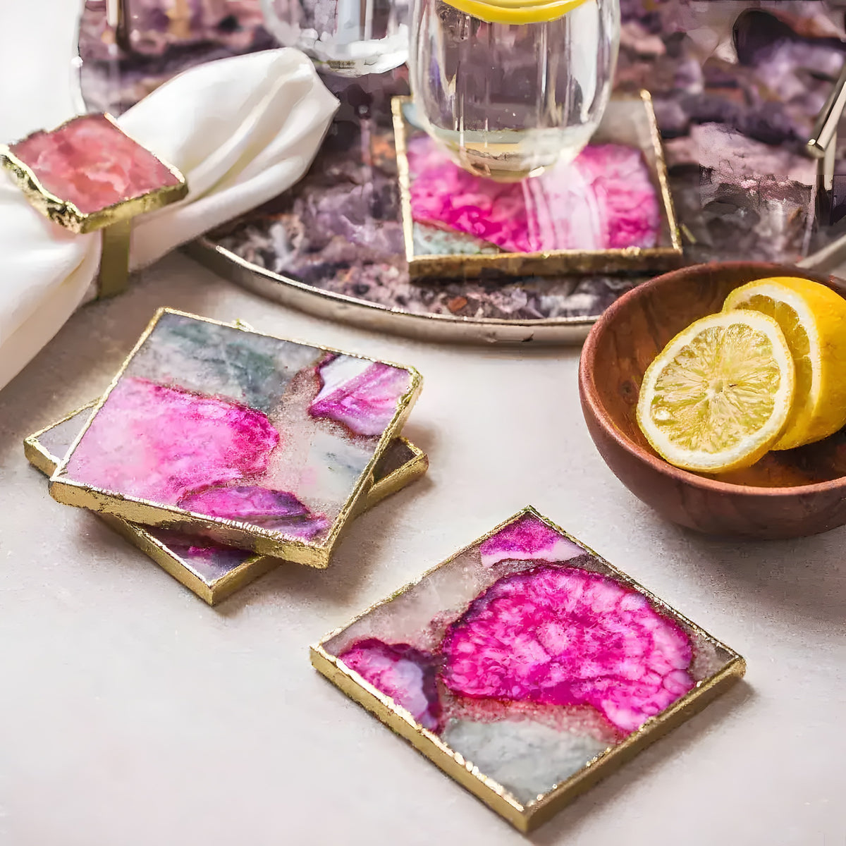 Square pink agate coasters with gold trim arranged on a table next to a glass and lemon slices