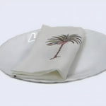 Cream cotton hand block napkin with embroidered palm tree motif, folded on white ceramic plate