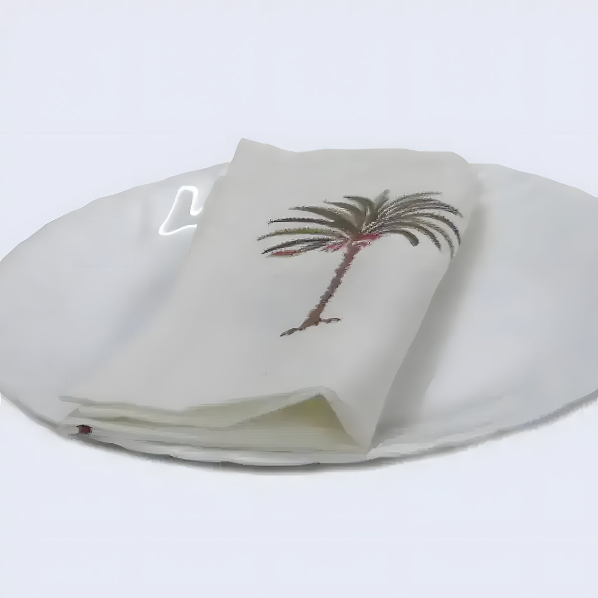 Cream cotton hand block napkin with embroidered palm tree motif, folded on white ceramic plate