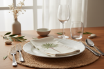 A styled table setting with a white ceramic plate, matching napkin featuring a green palm tree print, woven placemat, silver cutlery, a wine glass, and a clear tumbler on a wooden table.