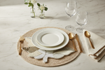 A styled table setting on a white marble surface featuring a woven round placemat, white textured ceramic plates, gold-toned flatware, a cream linen napkin, and two clear wine glasses.