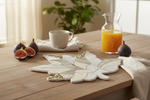 A decorative white and gold beaded leaf-shaped placemat on a wooden table, styled with a white mug, an orange juice carafe, and fresh figs.