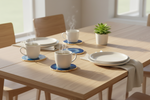 A styled dining table setup with four white ceramic mugs on blue coasters, white plates, beige napkins, and a small potted succulent on a light wood table in a bright room.