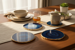 Four round agate-pattern coasters with gold-edged borders arranged on a wooden dining table alongside a beige ceramic mug, a glass of amber liquid, and other tableware in a softly lit interior setting.