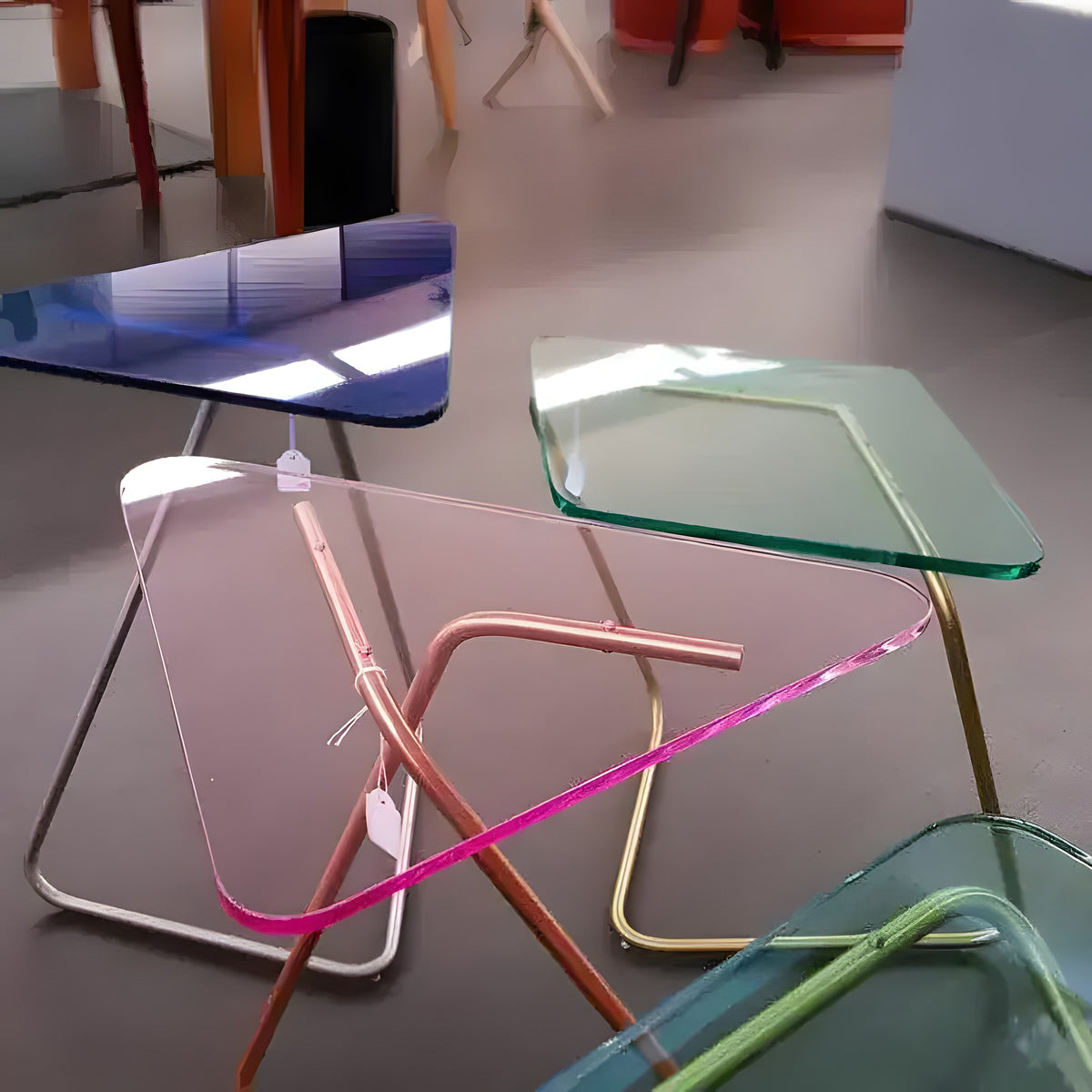 Multiple colorful triangle crystal tables with glass tops in pink, green and blue, on wire metal legs in a showroom