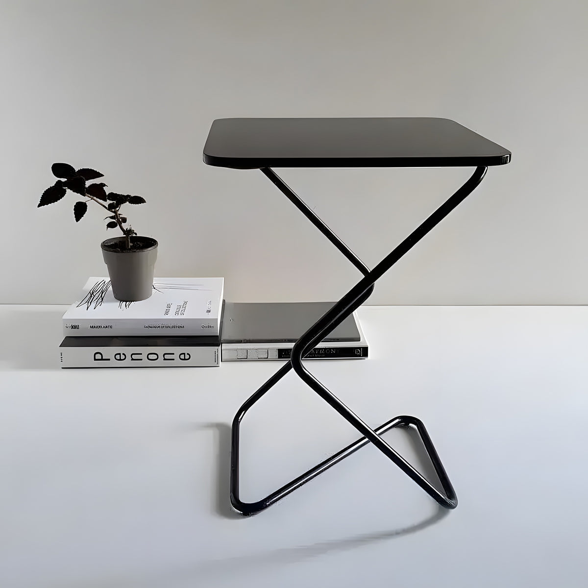 Black square side table with zigzag metal frame, styled next to stacked books and small potted plant on white surface