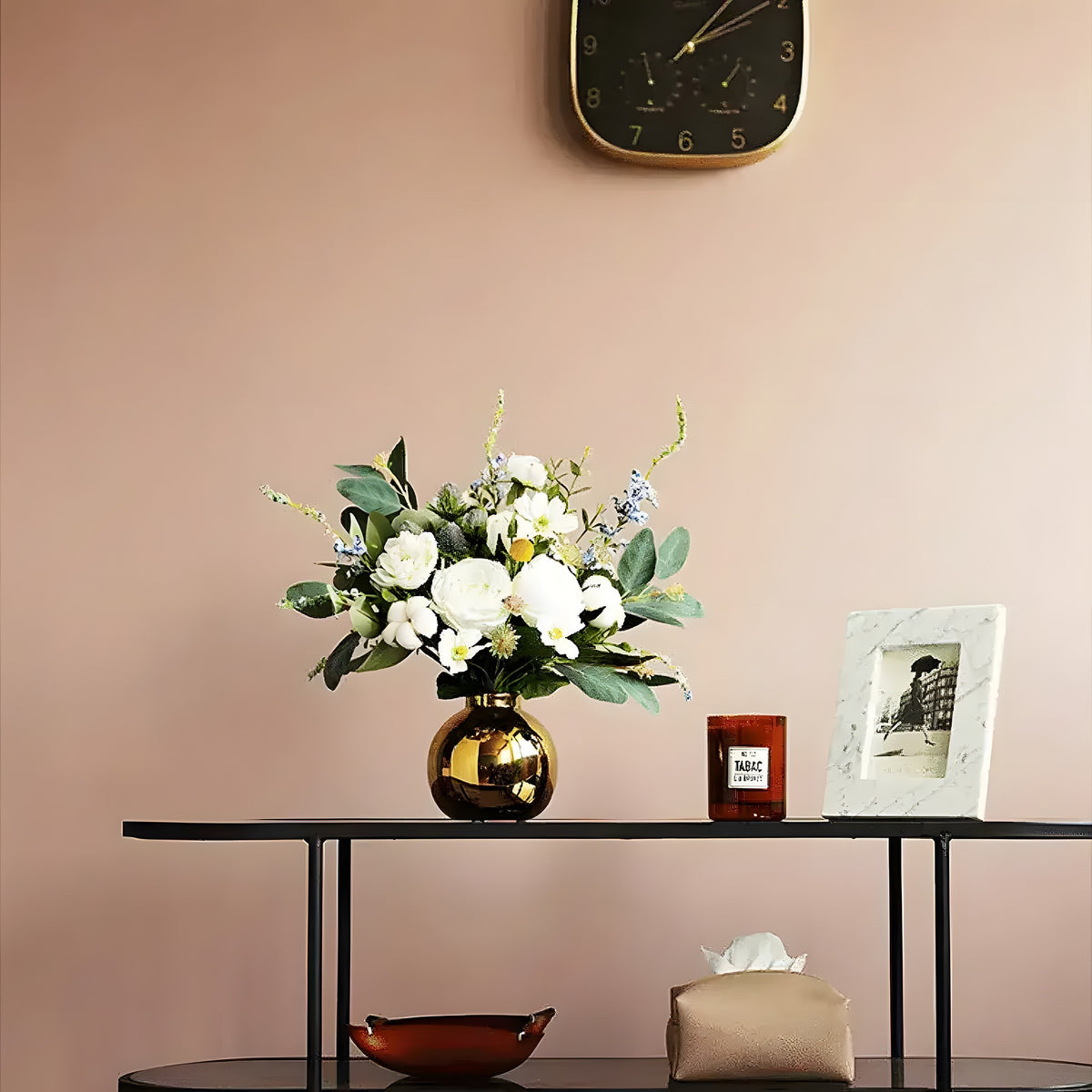 White cotton rose floral arrangement in round gold vase on black console with wall clock and marble frame