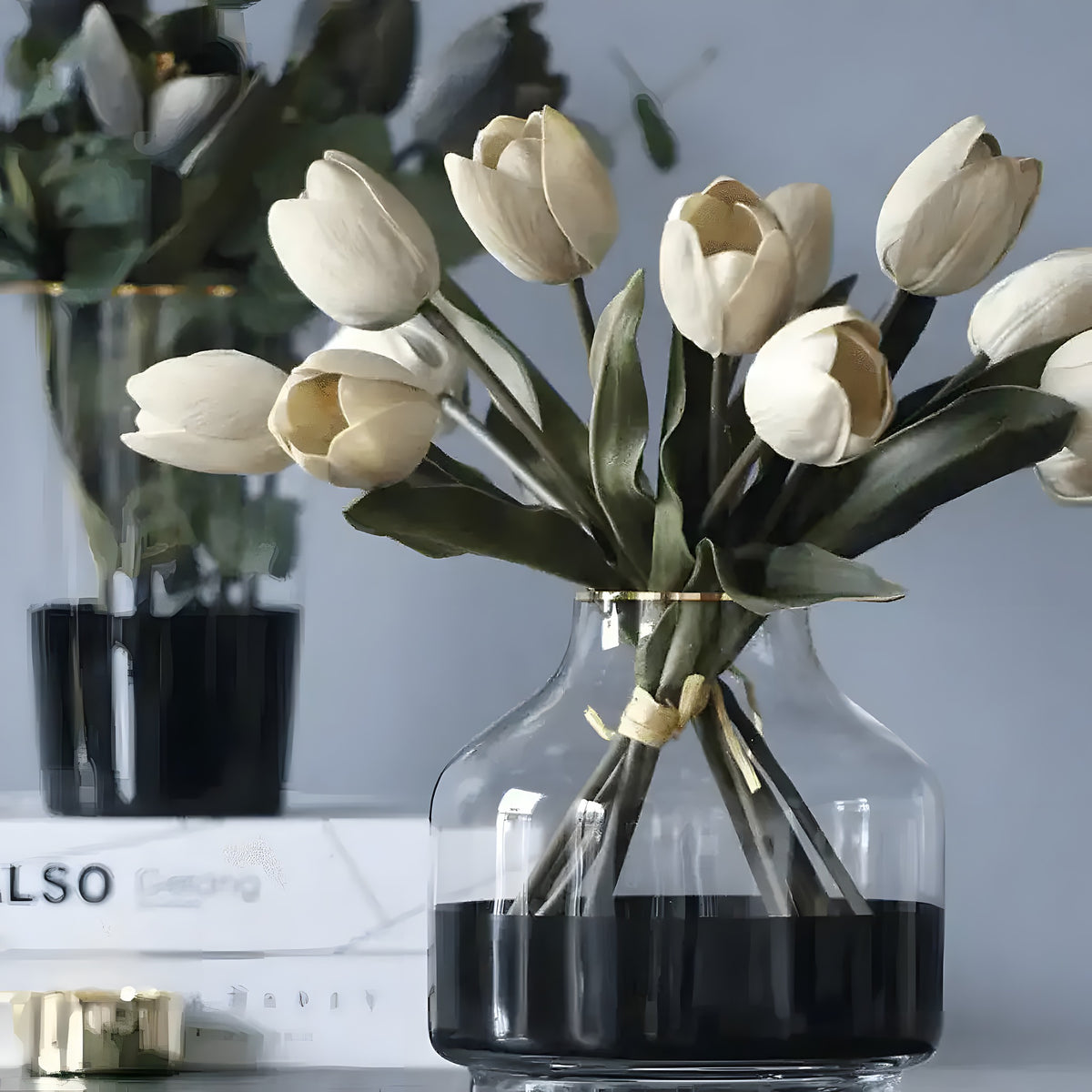Cream artificial real-touch tulips in squat glass vase with dark-tinted base on desk beside stacked books