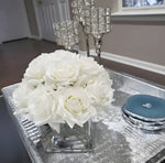 Dense faux white silk rose arrangement in square glass vase on silver tray, crystal candelabra in background